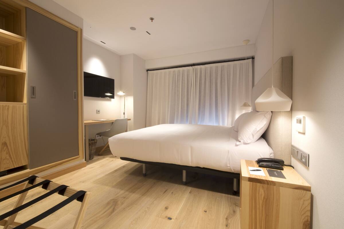 Une chambre d’hôtel moderne avec un grand lit, une table de chevet avec une lampe, un téléviseur fixé au mur et une armoire en bois.