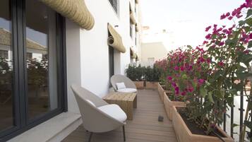 A balcony with wooden furniture, a white wall with woven decorations, and potted plants with pink flowers.