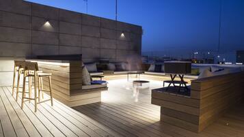 Terraza exterior con mobiliario de madera, zona de bar y vistas a la ciudad al atardecer.