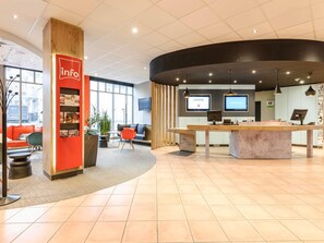 Ett modernt receptionsområde med en receptionsdisk i trä, en sittgrupp med orange och gröna stolar samt ett stort fönster med utsikt över gatan.