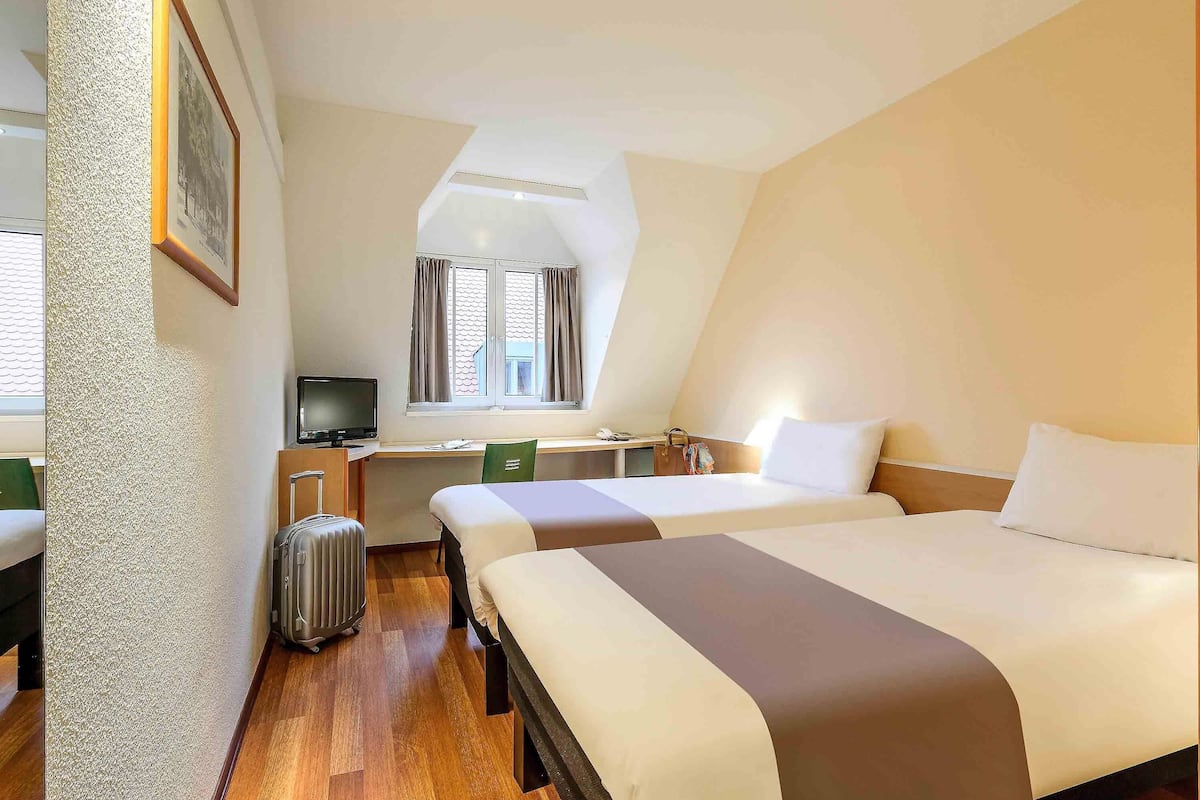 A hotel room with two beds, a desk, a chair, a television, and a suitcase.