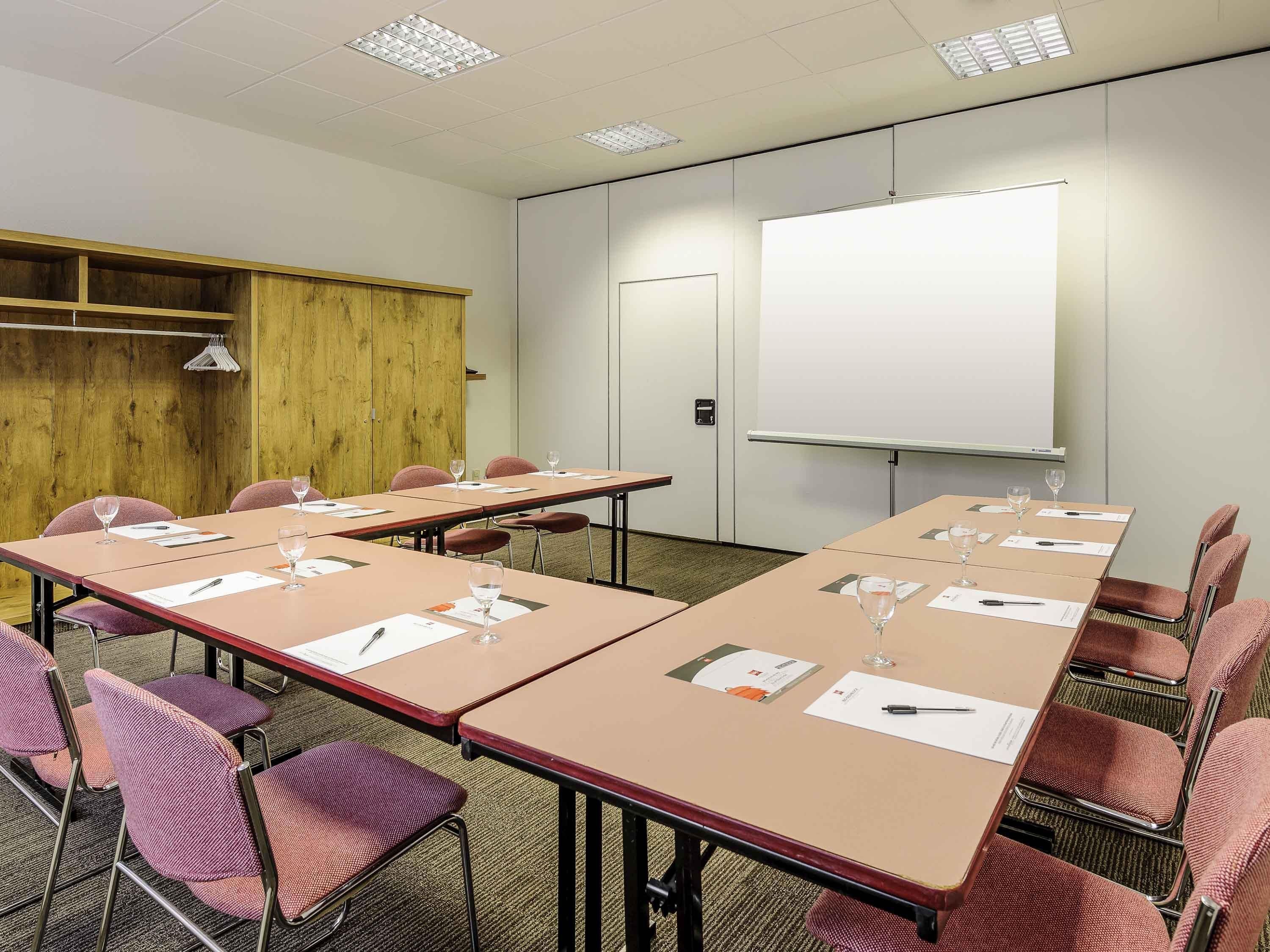 Una sala conferenze con lunghe tavolate, sedie, una lavagna e un armadio in legno.