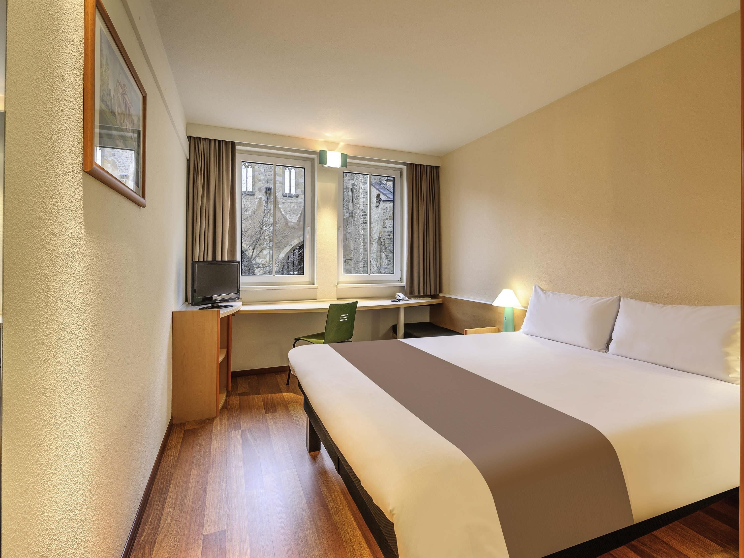 A hotel room with a bed, desk, chair, and a window with curtains.