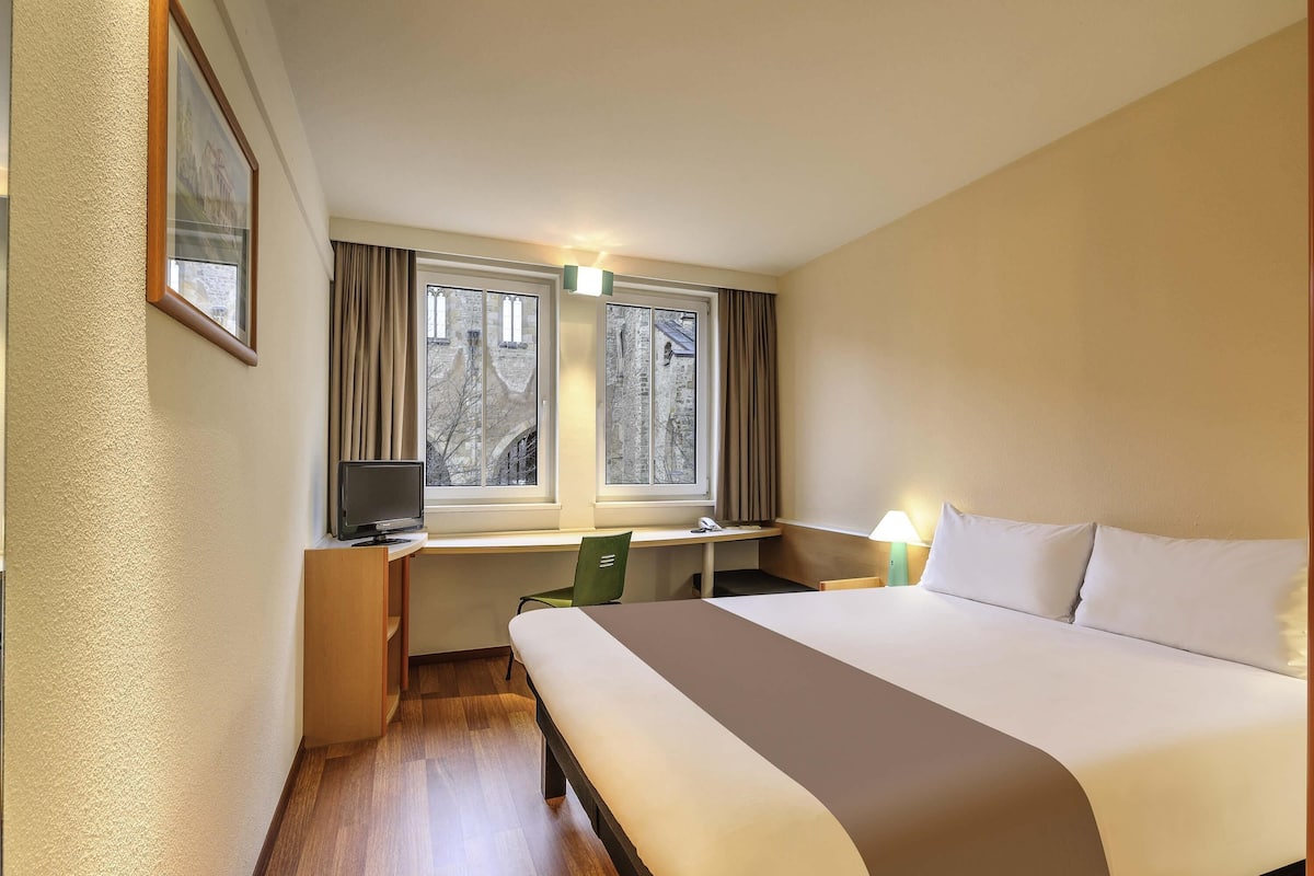 A hotel room with a bed, desk, chair, and a window with curtains.