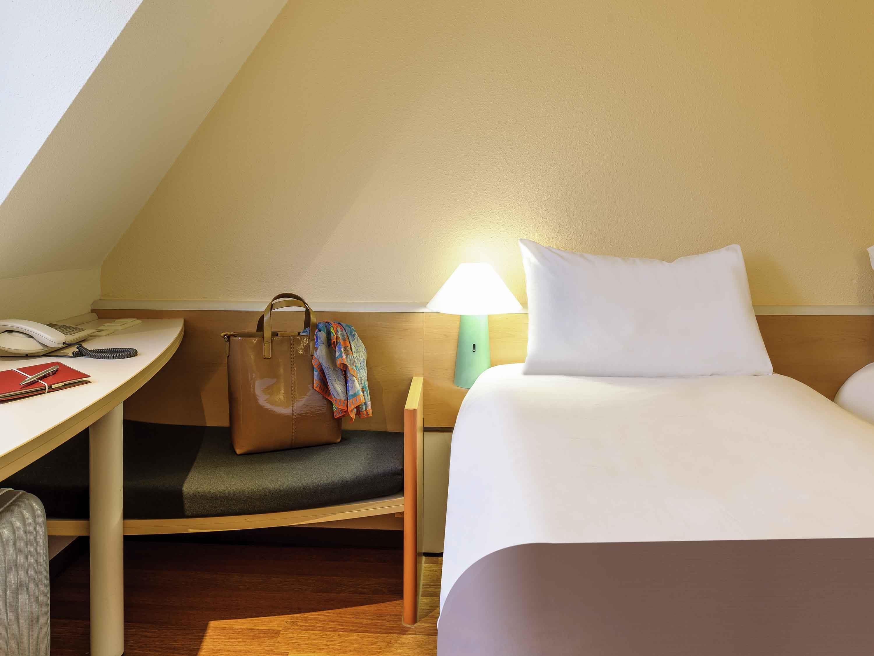 A hotel room with a desk, a telephone, a lamp, a bed, and a suitcase.
