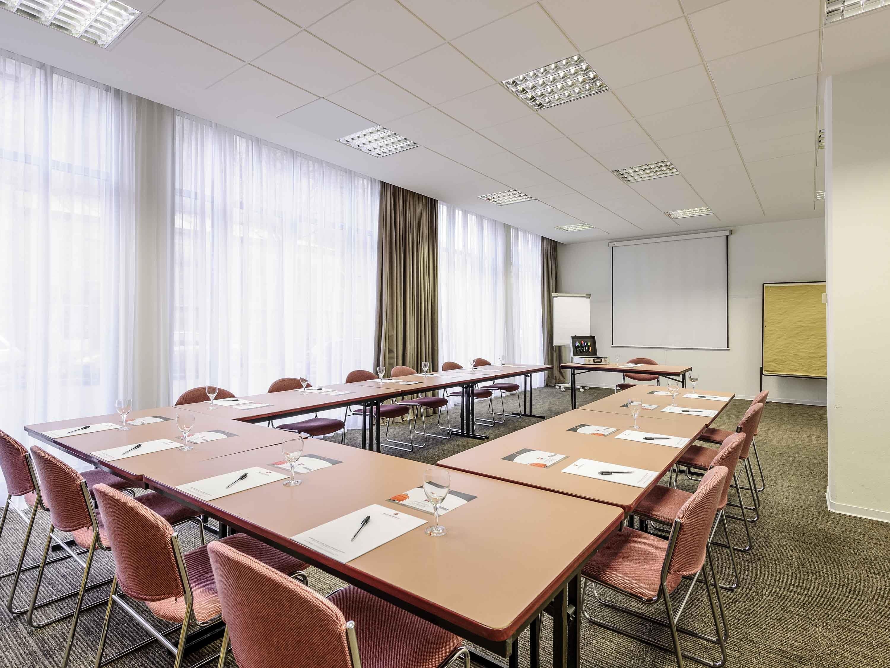 Ein Konferenzraum mit langen Tischen, Stühlen, einer Projektionsleinwand und einem großen Fenster mit Vorhängen.