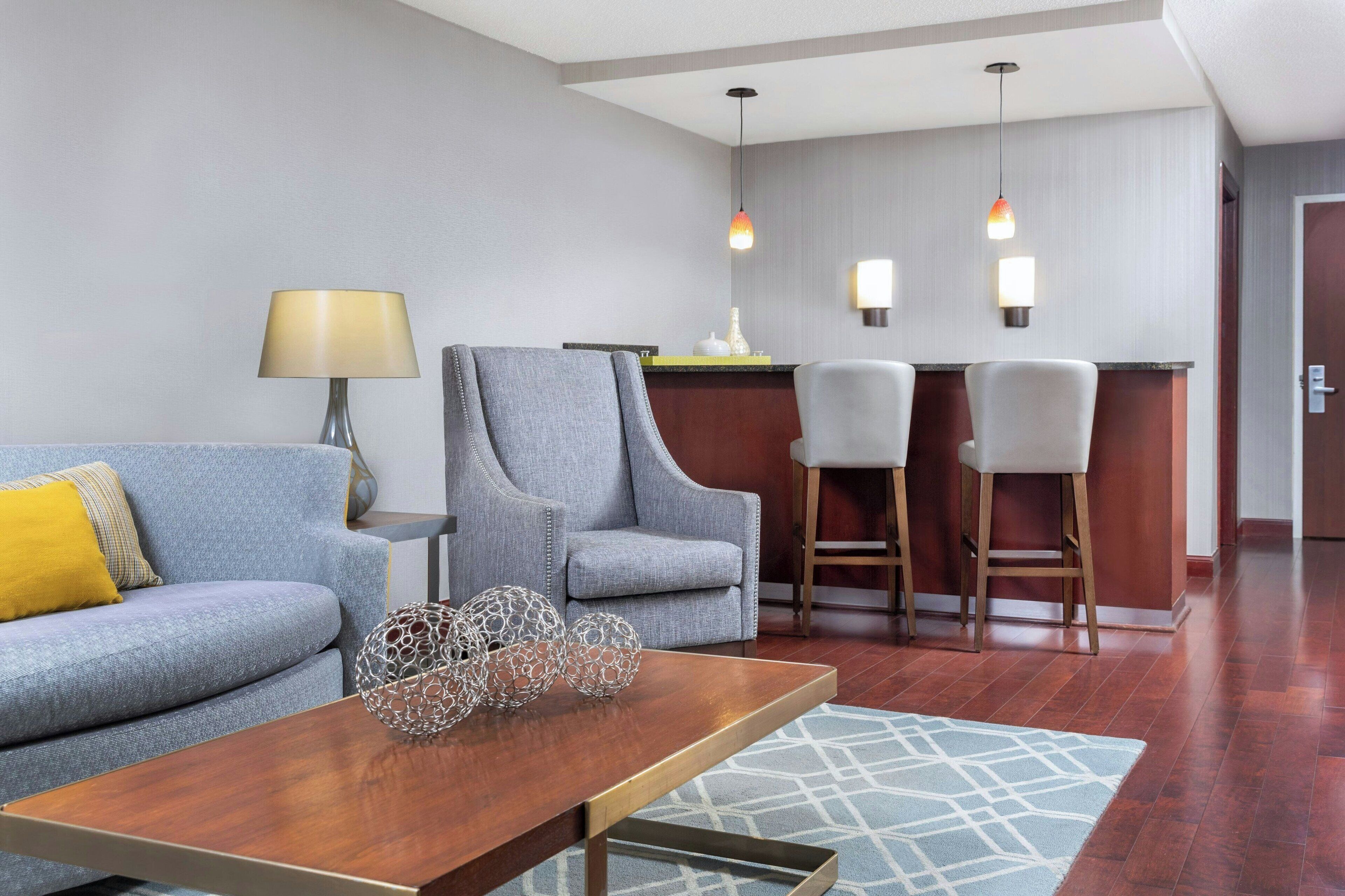 A modern hotel room with a grey sofa, a wooden coffee table, and a bar area with two bar stools.