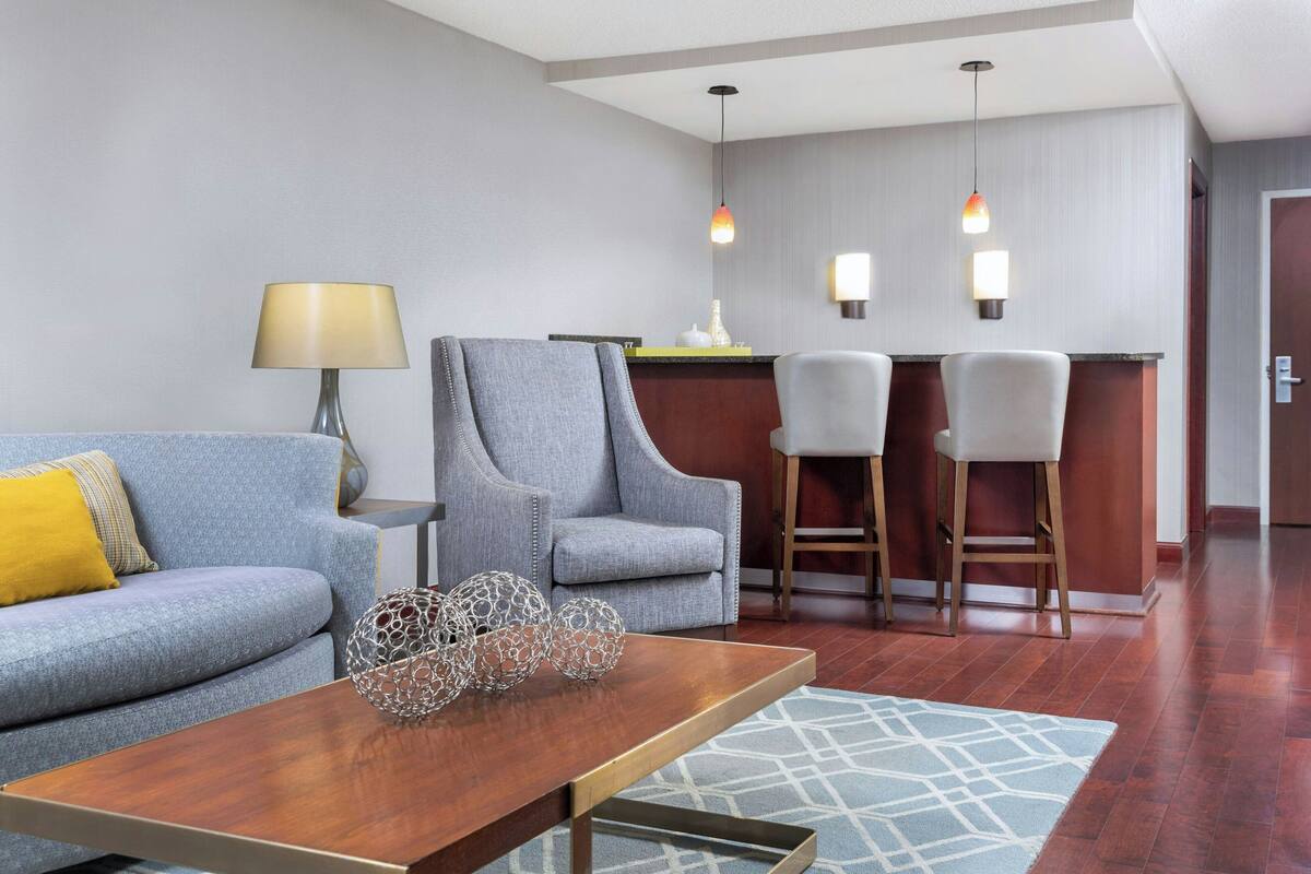 A modern hotel room with a grey sofa, a wooden coffee table, and a bar area with two bar stools.