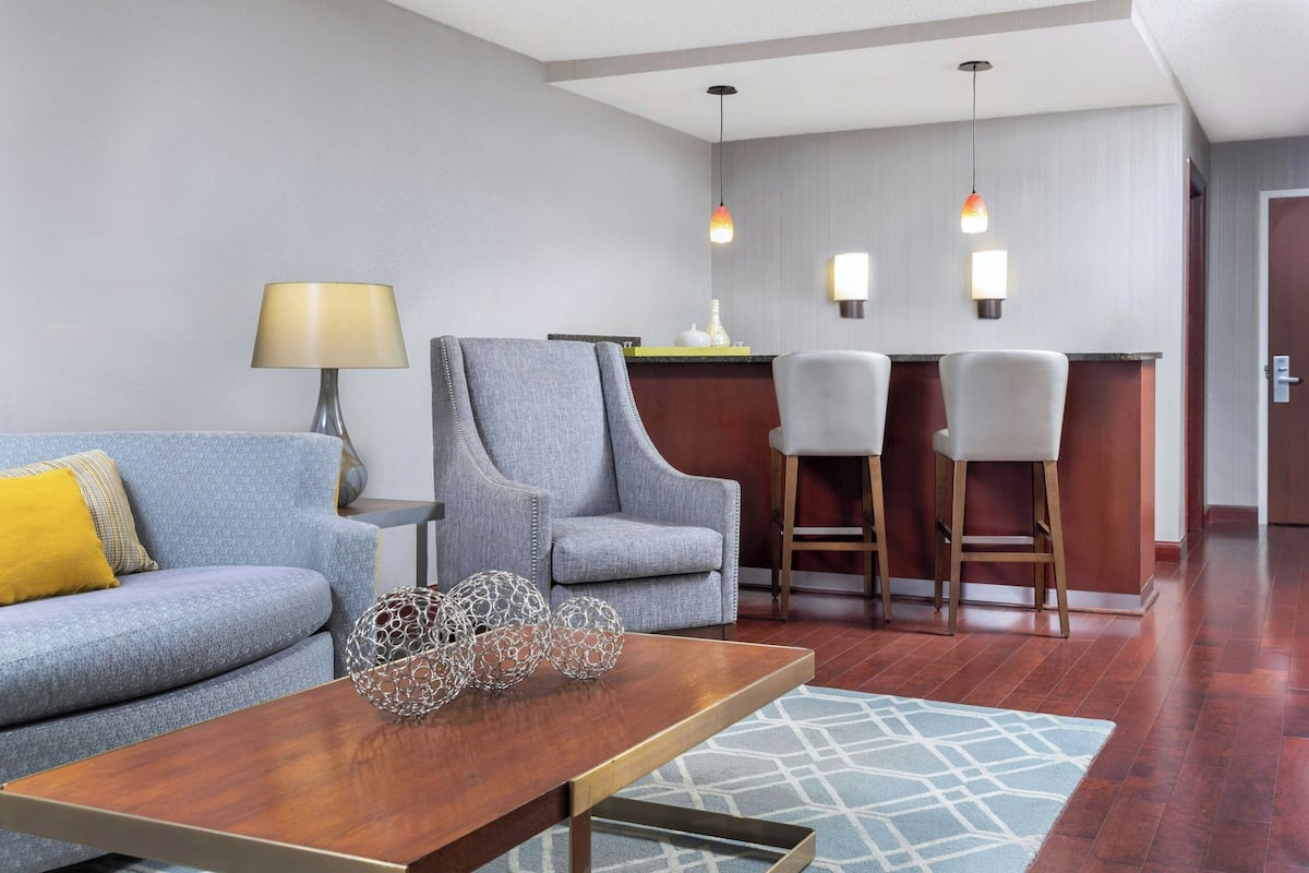 A modern hotel room with a grey sofa, a wooden coffee table, and a bar area with two bar stools.
