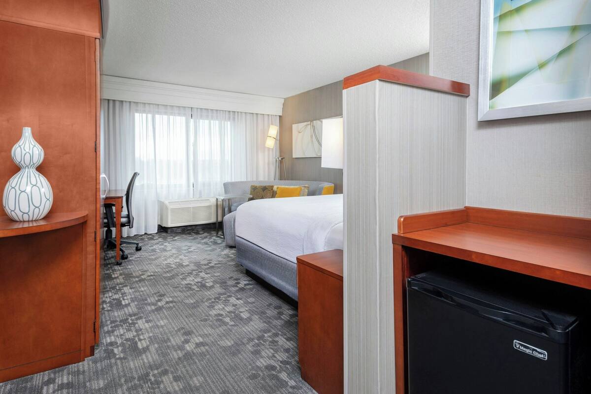 A hotel room with a bed, a desk, a chair, a minibar, and a trash bin.
