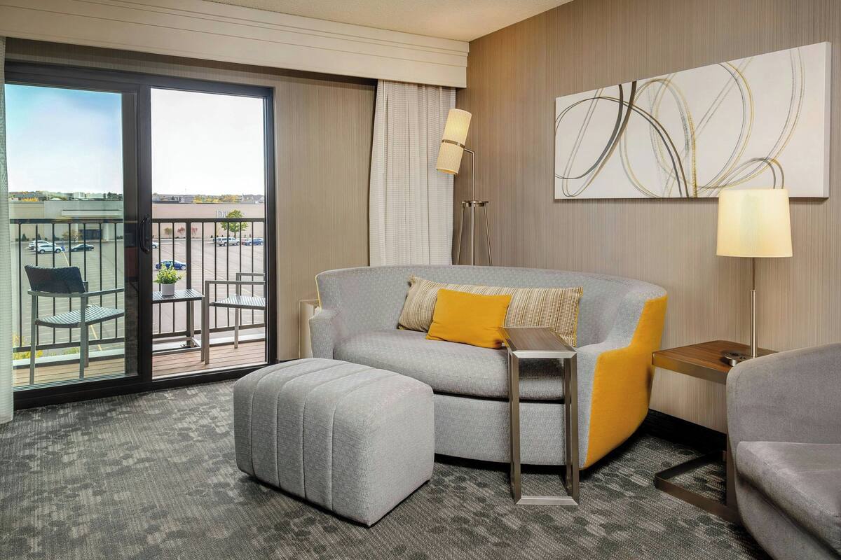 A modern hotel room with a grey sofa, a matching ottoman, a small wooden side table, and a floor lamp. A large window offers a view of a parking area and a building across the street.