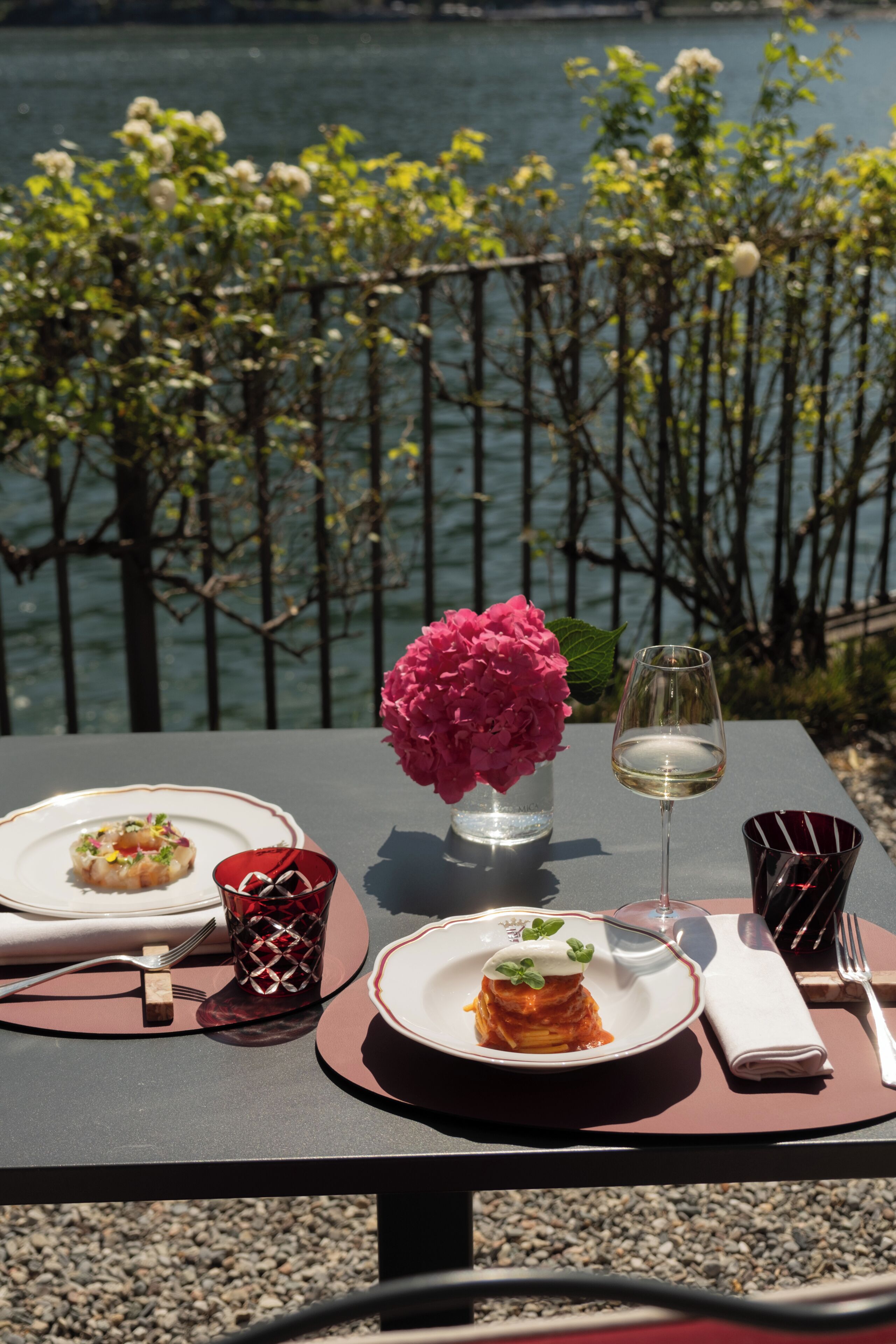 Un tavolo apparecchiato con piatti di cibo, un bicchiere di vino e un vaso di fiori, con vista su un'area d'acqua.