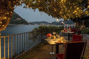 Un área de comedor al aire libre con una mesa dispuesta para dos, con vista a un cuerpo de agua y un paisaje urbano.
