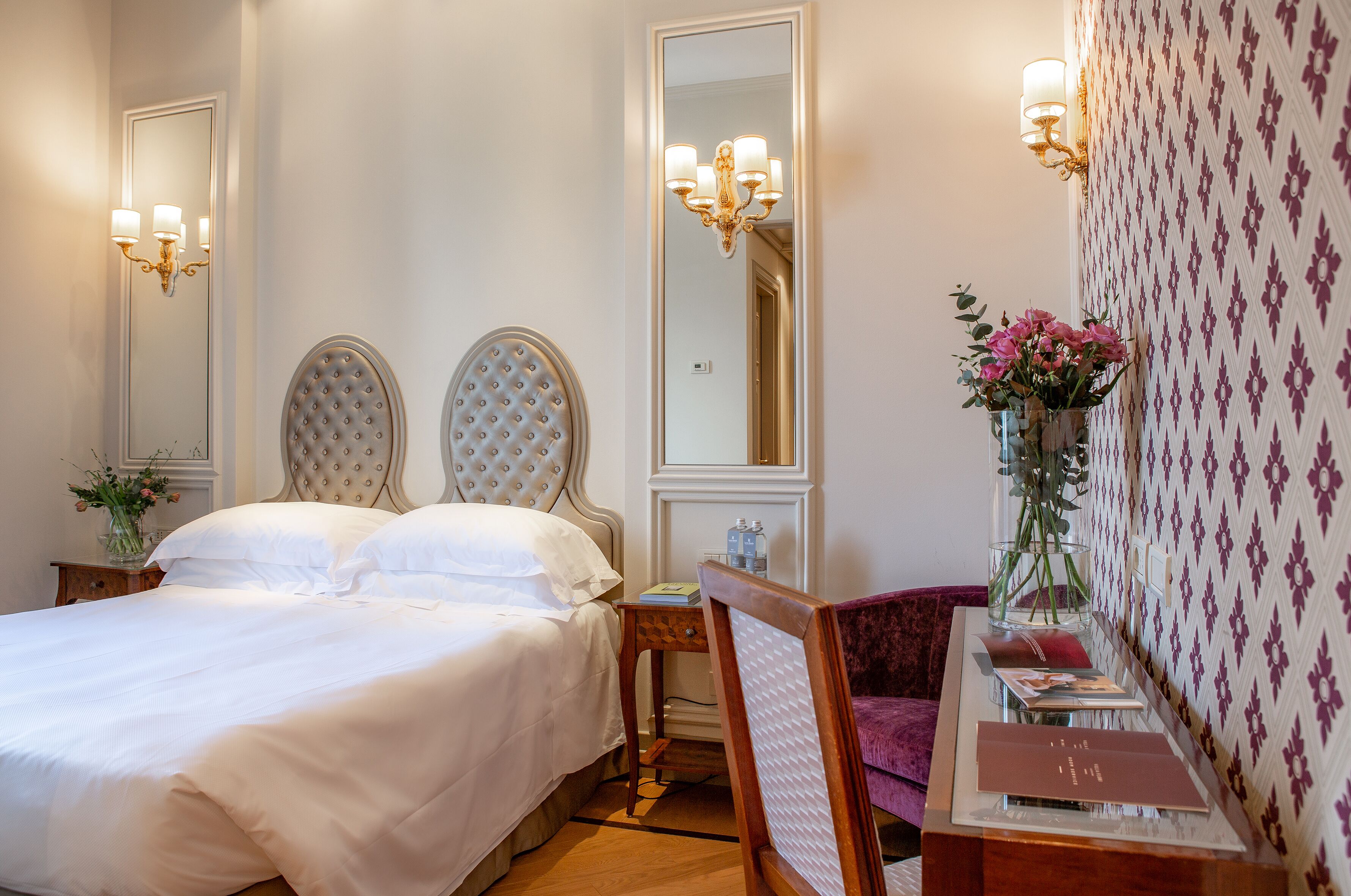 A hotel room with a large bed, a vanity with a mirror, a chair, and a table with a vase of flowers.