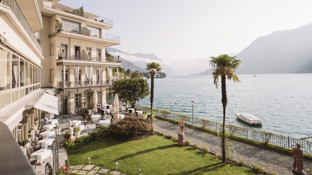 A hotel with outdoor seating overlooking a lake, a statue, and mountains in the background.