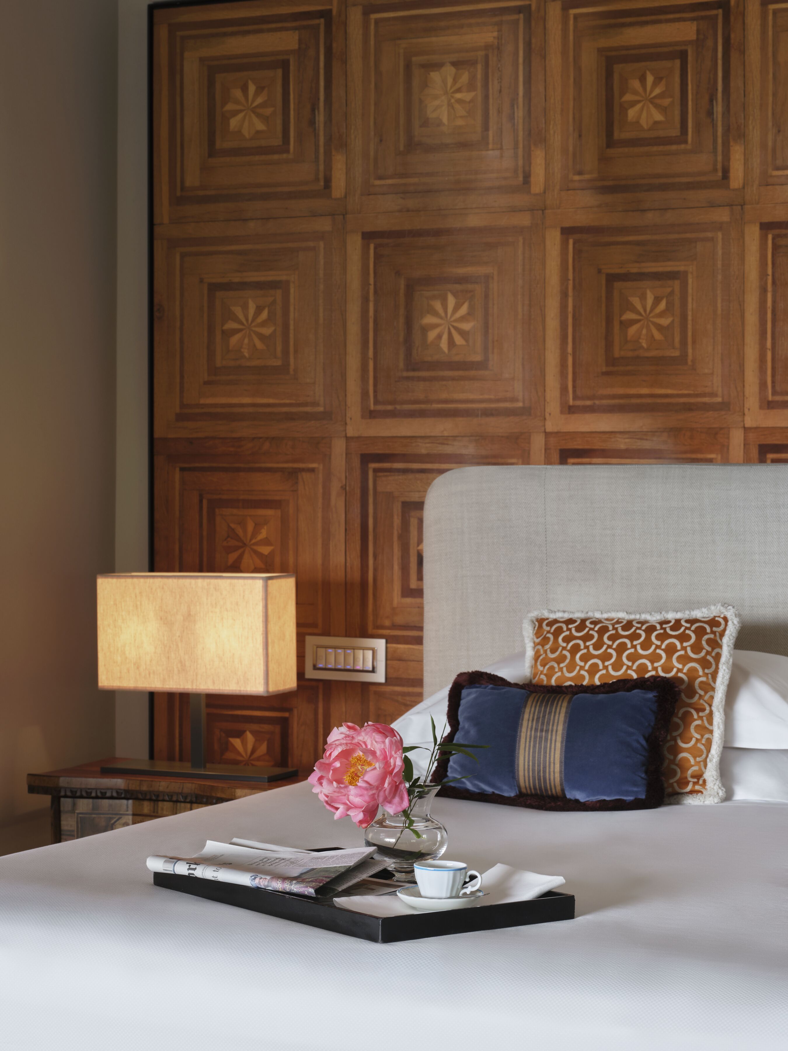 Un letto ben rifatto con una testiera decorativa, una lampada da comodino, un vaso con un fiore rosa e un vassoio con una tazza e alcune riviste.