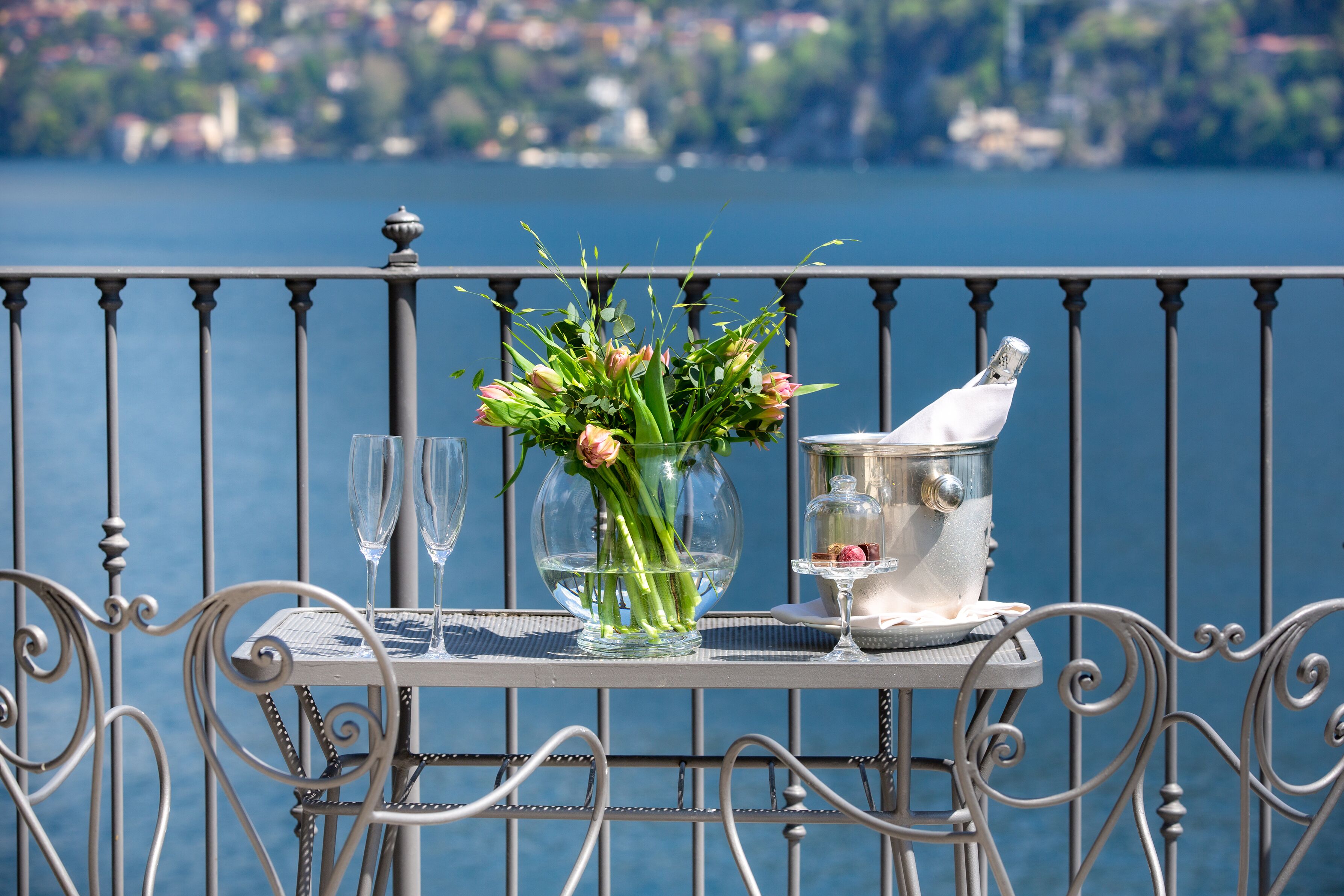 A table with a vase of flowers, two glasses, and a bottle of champagne on a balcony with a view of a lake and buildings.