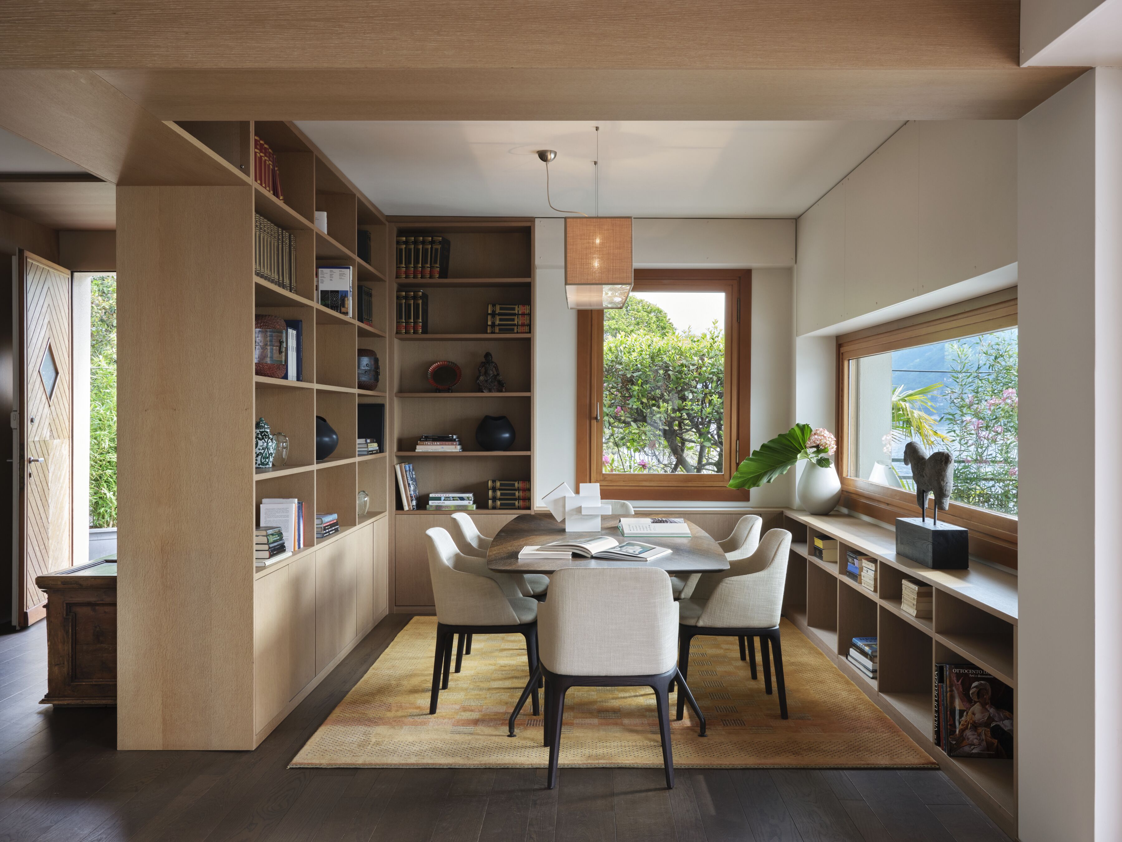 Una biblioteca moderna con zona de comedor, estanterías y un amplio ventanal con vista a árboles.