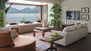 Una sala de estar moderna con un amplio ventanal con vistas a un lago y montañas, un sofá blanco, un sillón marrón y una mesa de centro con libros y limones.