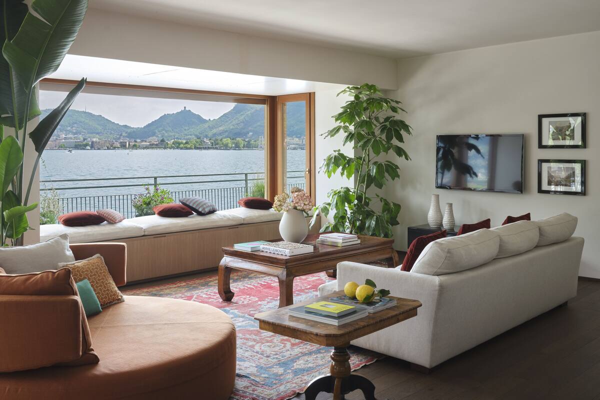 Una sala de estar moderna con un amplio ventanal con vistas a un lago y montañas, un sofá blanco, un sillón marrón y una mesa de centro con libros y limones.