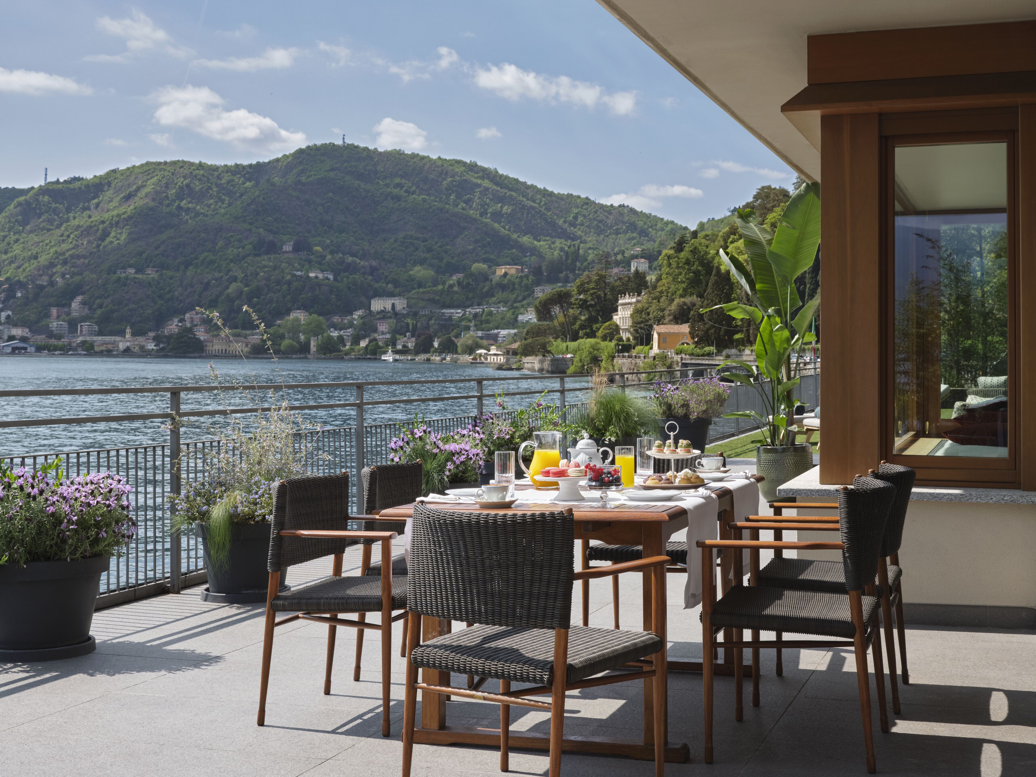Una terraza con sillas de mimbre, una mesa preparada para el desayuno y vistas a un lago y colinas.