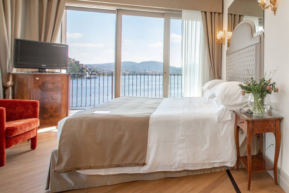 Habitación de hotel con una cama grande, una silla roja, un televisor sobre un mueble de madera y vistas al mar a través de amplios ventanales.