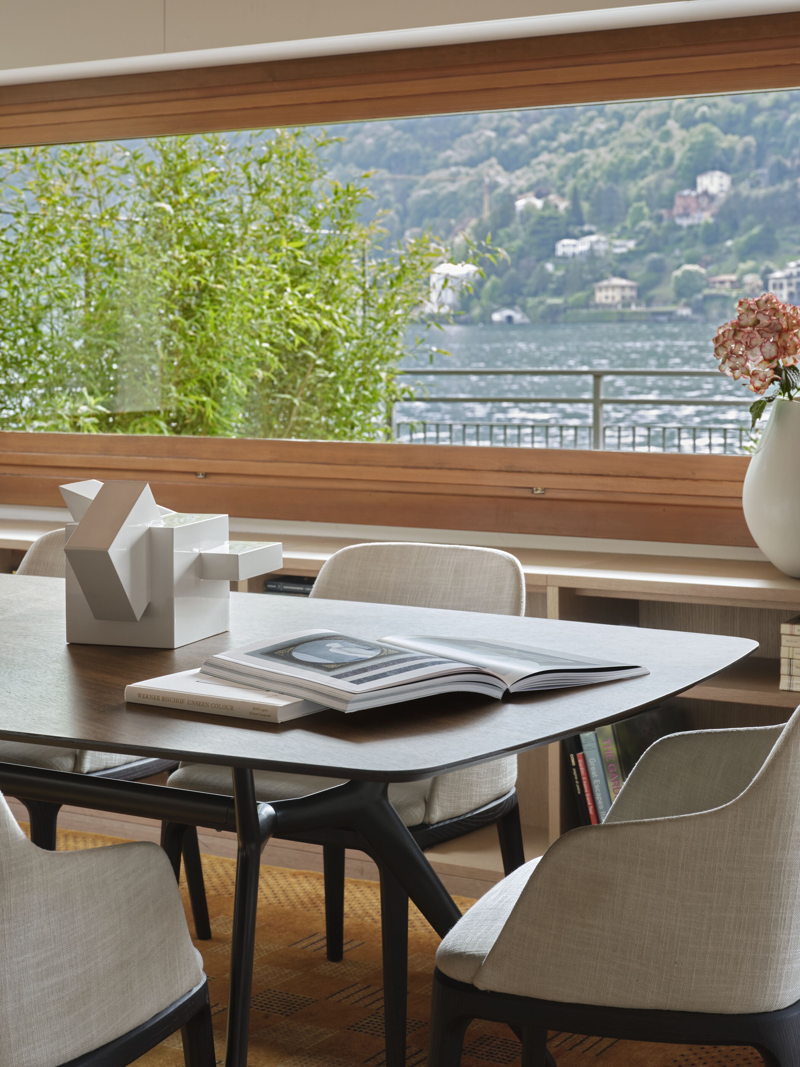 Un comedor moderno con vistas a un lago y colinas. Hay una mesa con una revista, un jarrón con flores y una escultura blanca.