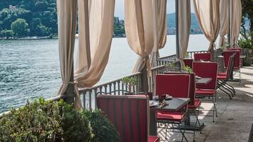 Una terraza con sillas rojas y cortinas color crema, con vistas a un lago y una montaña al fondo.
