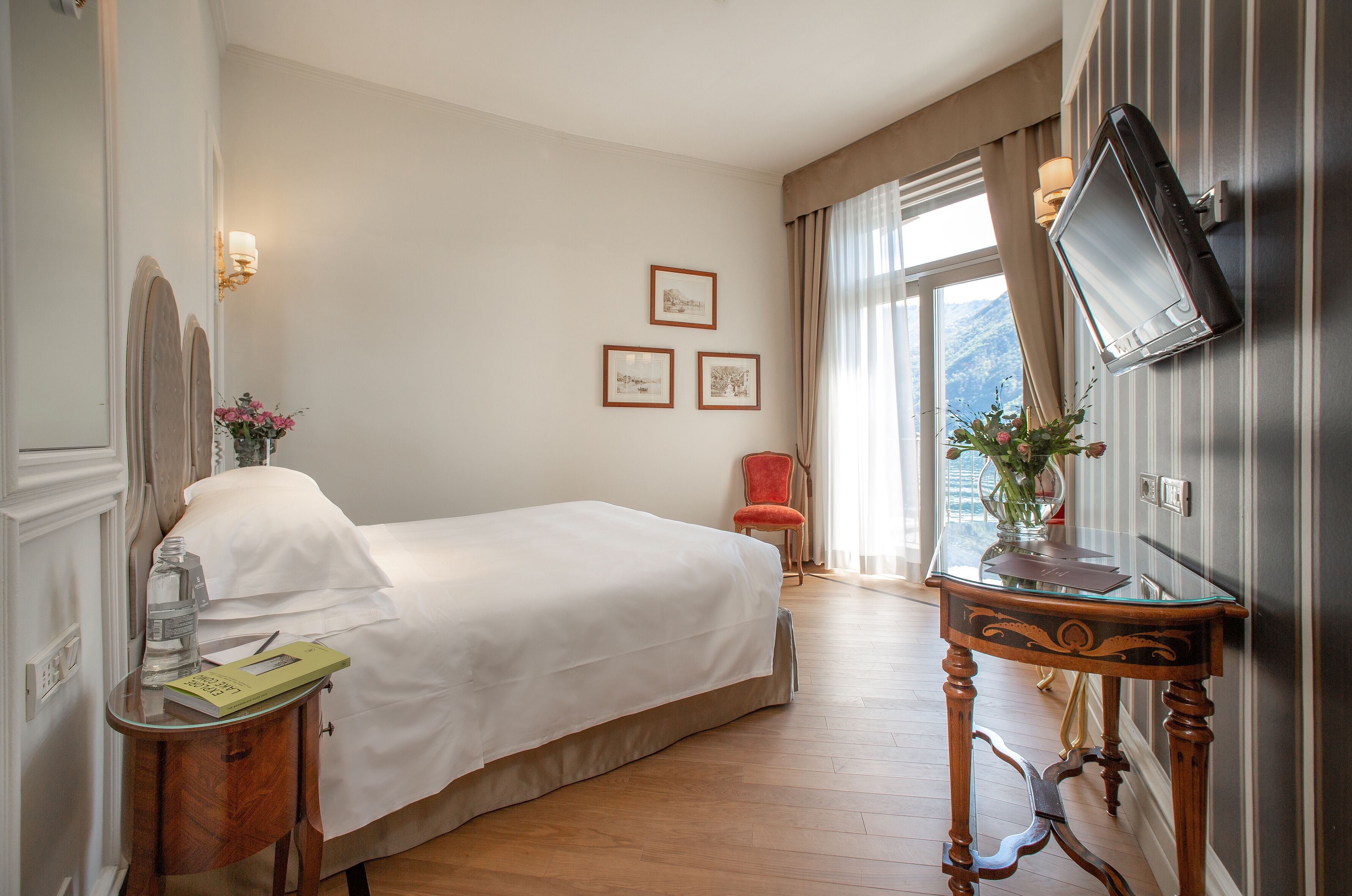 Ein Hotelzimmer mit einem Bett, einem Nachttisch mit Buch und Flasche, einem Sessel, einem an der Wand befestigten Fernseher und gerahmten Bildern.