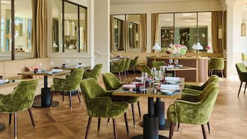 A spacious hotel dining area with green velvet chairs, a long table set for dining, and a bar area in the background.