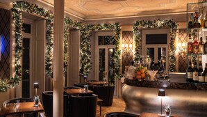 A warmly lit interior with Christmas decorations, including garlands and a Christmas tree. There are tables and chairs arranged for dining, and a bar area with bottles and glasses.