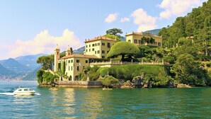 A large, historic building with multiple stories and a tower, situated on a lakeside with lush greenery and mountains in the background.