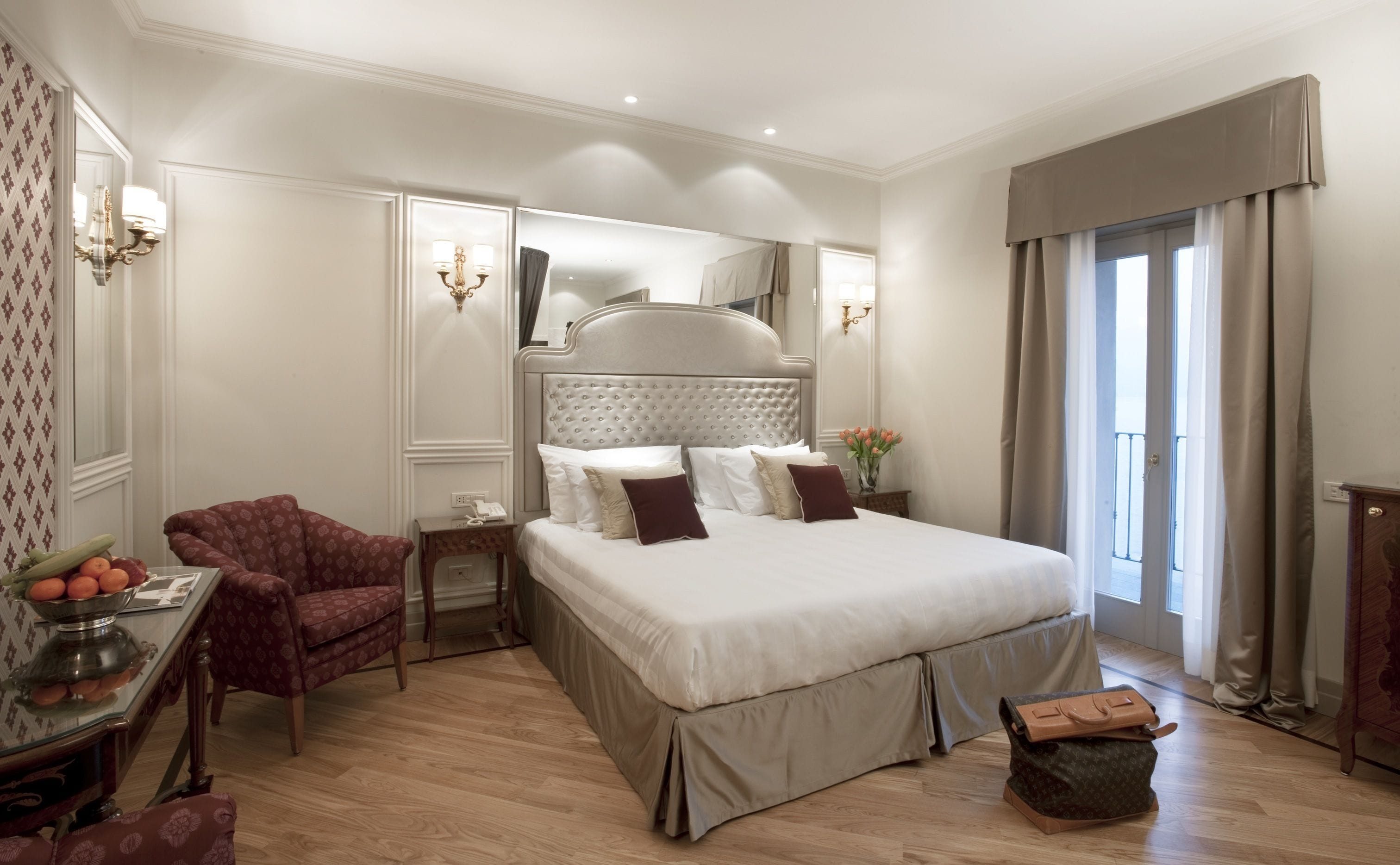 A spacious bedroom with a large bed, a sitting area with a chair, a small table with a fruit bowl, and a mirror.