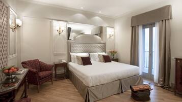 A spacious bedroom with a large bed, a sitting area with a chair, a small table with a fruit bowl, and a mirror.