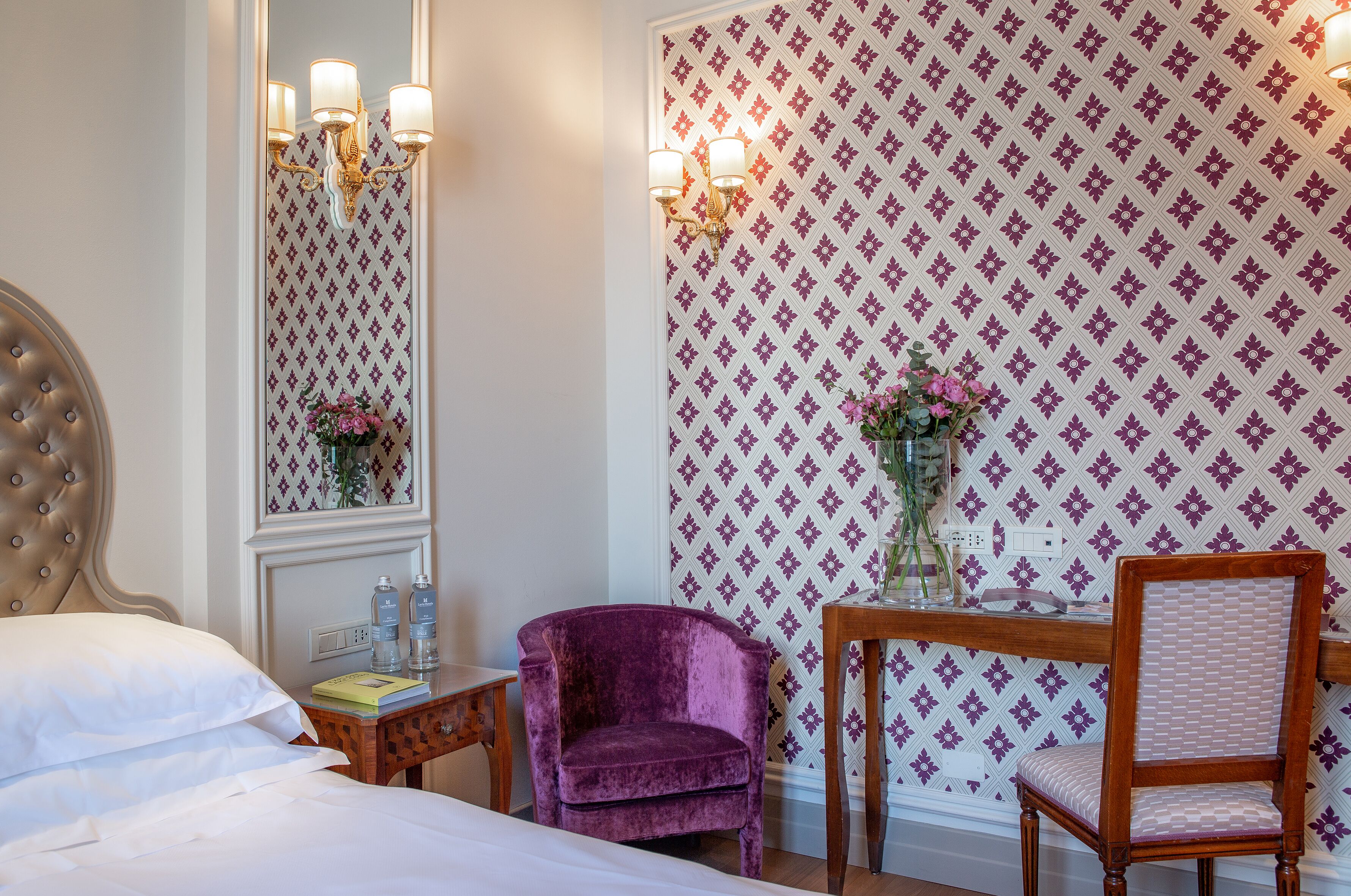 A hotel room with a bed, a vanity with a mirror, a purple chair, and a wooden table with a vase of flowers.