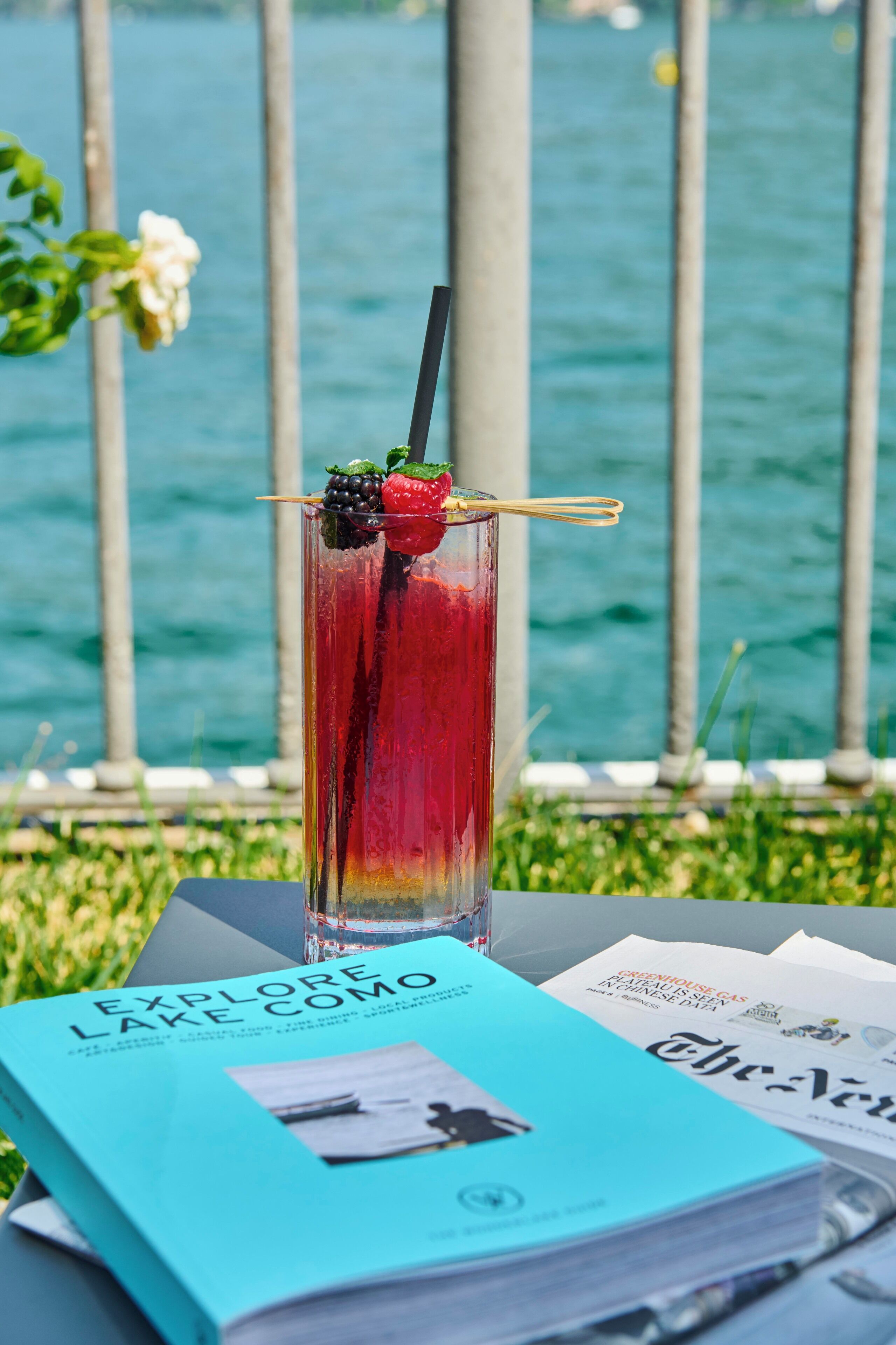 A tall glass with a red drink, garnished with a strawberry and blackberry, placed on a table with a book titled "Explore Lake Como" and a newspaper.