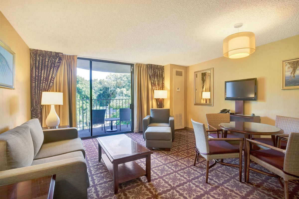 A hotel room with a sofa, armchair, ottoman, dining table, and chairs. There is a TV, a lamp, and a window with curtains.