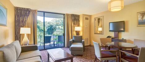A hotel room with a sofa, armchair, ottoman, dining table, and chairs. There is a TV, a lamp, and a window with curtains.