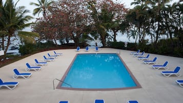 Una piscina con sillas reclinables azules dispuestas a su alrededor, rodeada de palmeras y vista al océano.