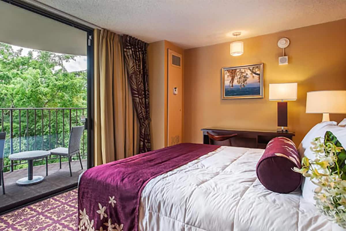 A hotel room with a bed, a desk, a chair, a balcony with a view of trees, and a painting on the wall.