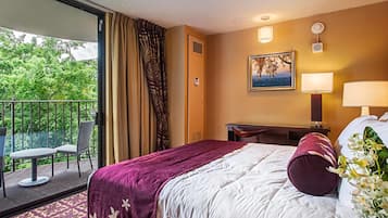 A hotel room with a bed, a desk, a chair, a balcony with a view of trees, and a painting on the wall.