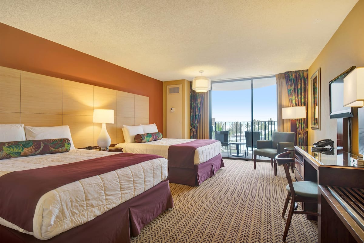 A hotel room with two beds, a desk, a chair, and a balcony with a view.