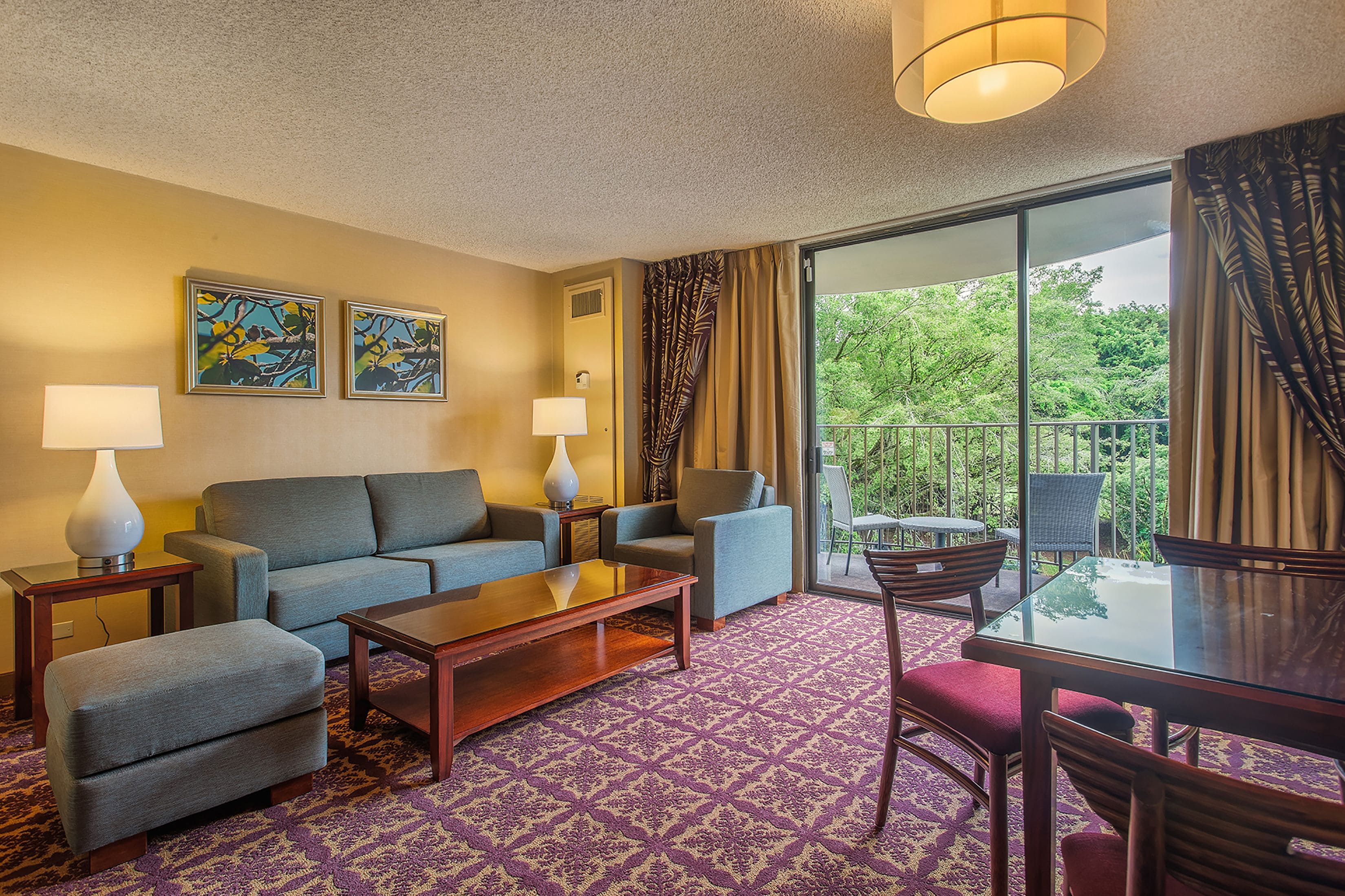 A hotel room with a sofa, armchairs, a coffee table, a dining table with chairs, and a balcony with a view of greenery.