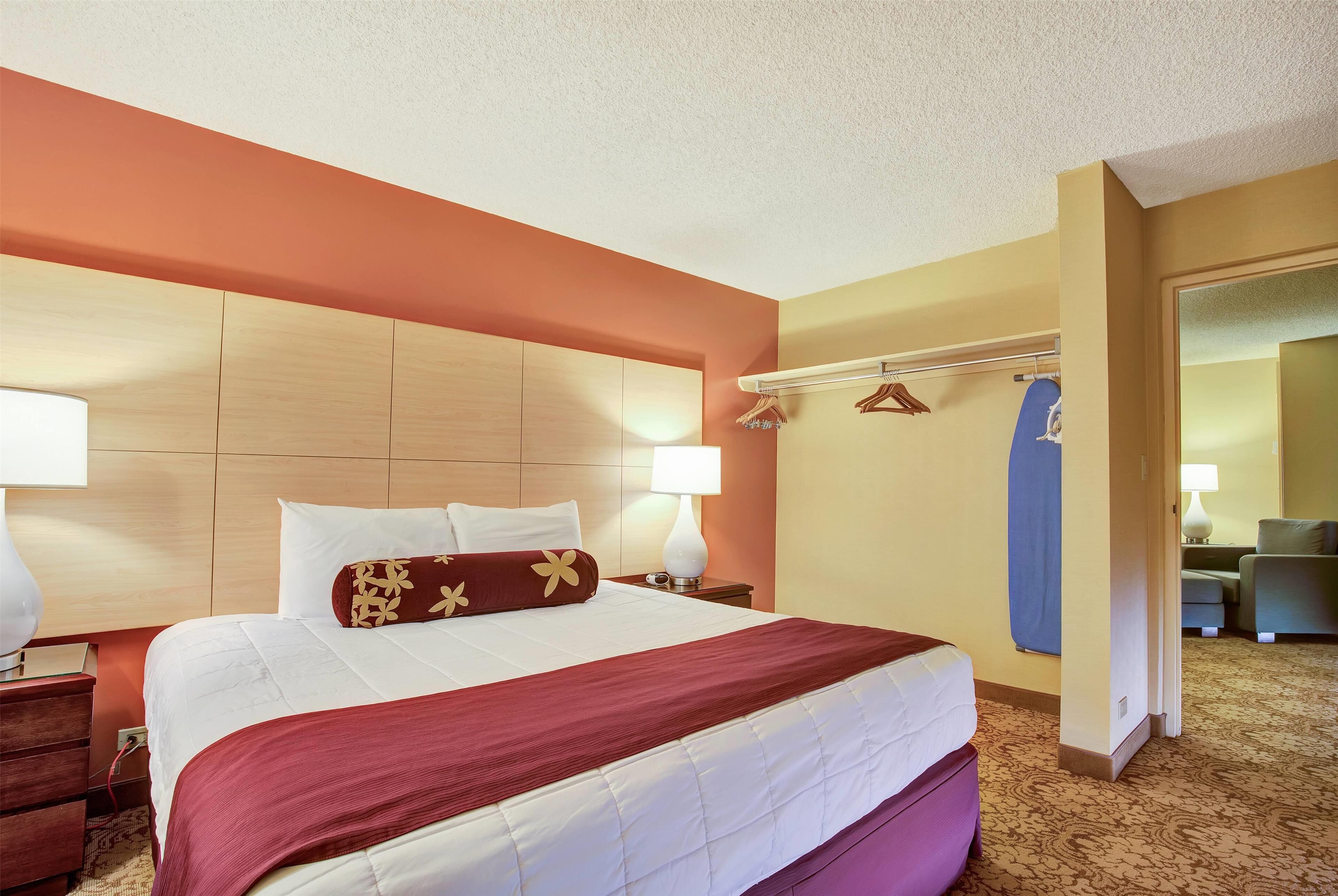 A hotel room with a large bed, wooden headboard, two bedside tables with lamps, a wardrobe, and a seating area with a sofa and chairs.