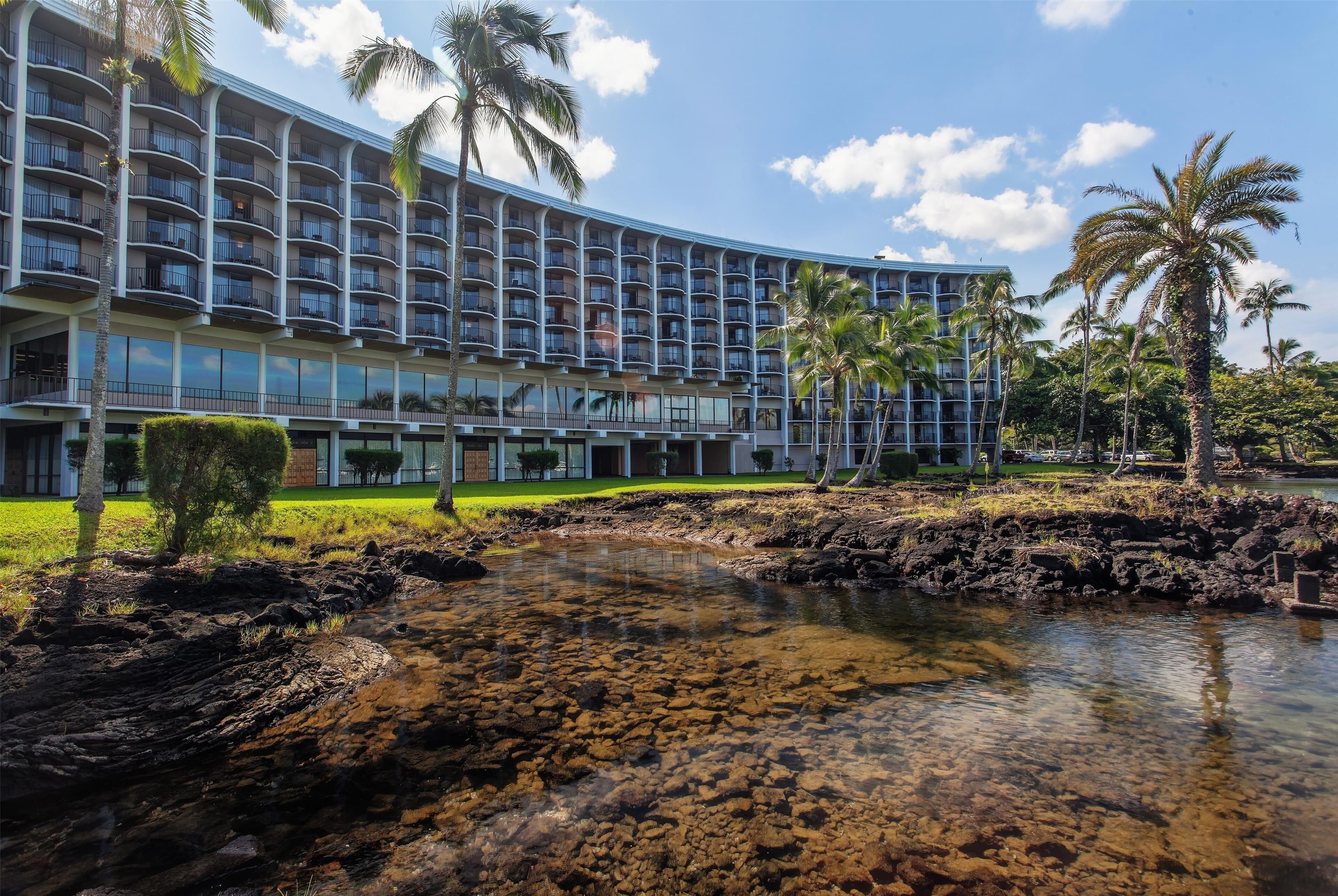 A hotel with multiple balconies, a clear water feature in front, and palm trees.