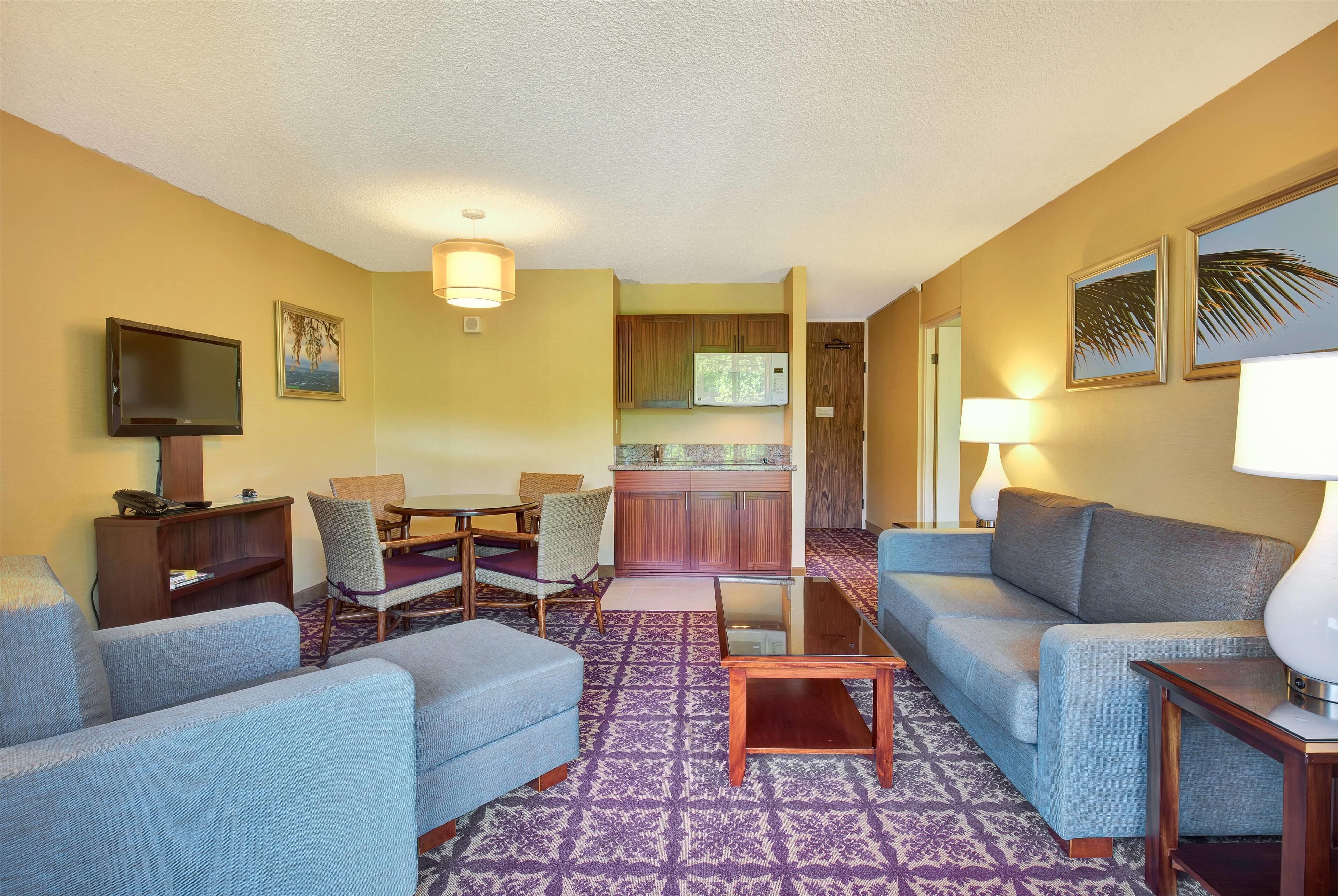 A living room with a sofa, chairs, a coffee table, and a TV. There is a kitchen area with a microwave and cabinets.
