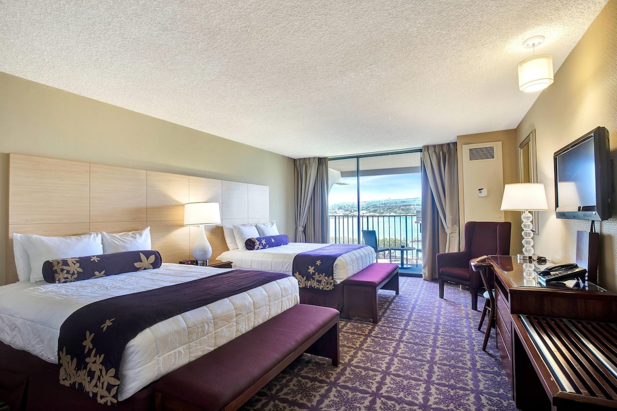 A hotel room with two beds, a desk, a chair, a bench, and a TV.