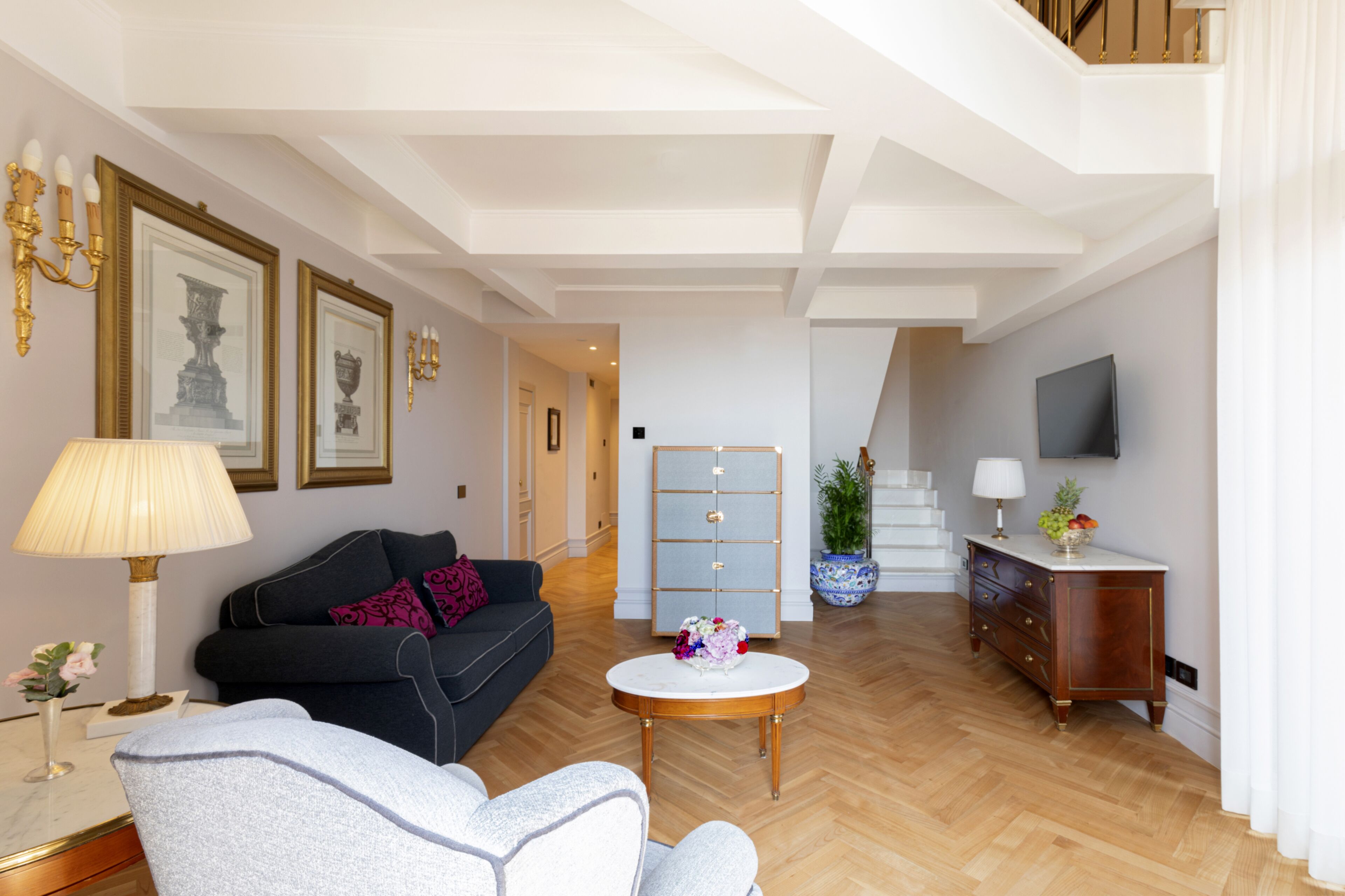 A spacious living room with a sofa, armchair, and coffee table. There is a television, a cabinet, and a staircase leading to an upper floor.
