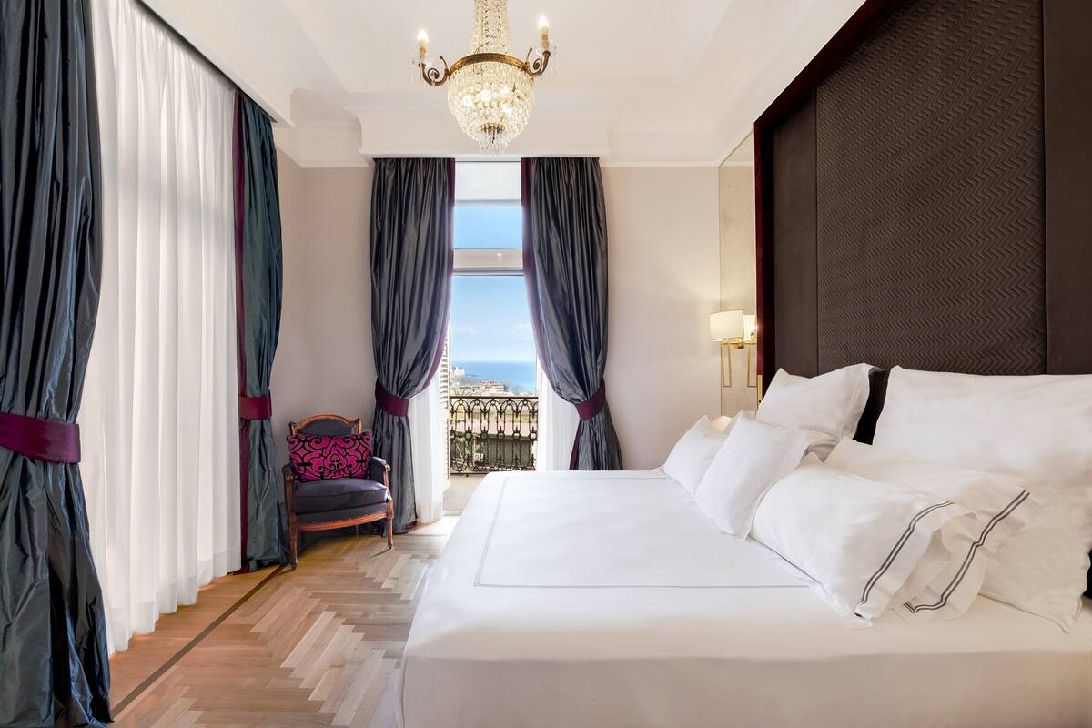 A hotel room with a large bed, a chandelier, a view of the city, and a balcony with curtains.