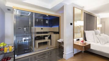 Habitación de hotel con ducha de vidrio, cama con ropa blanca y mesita de noche con lámpara.